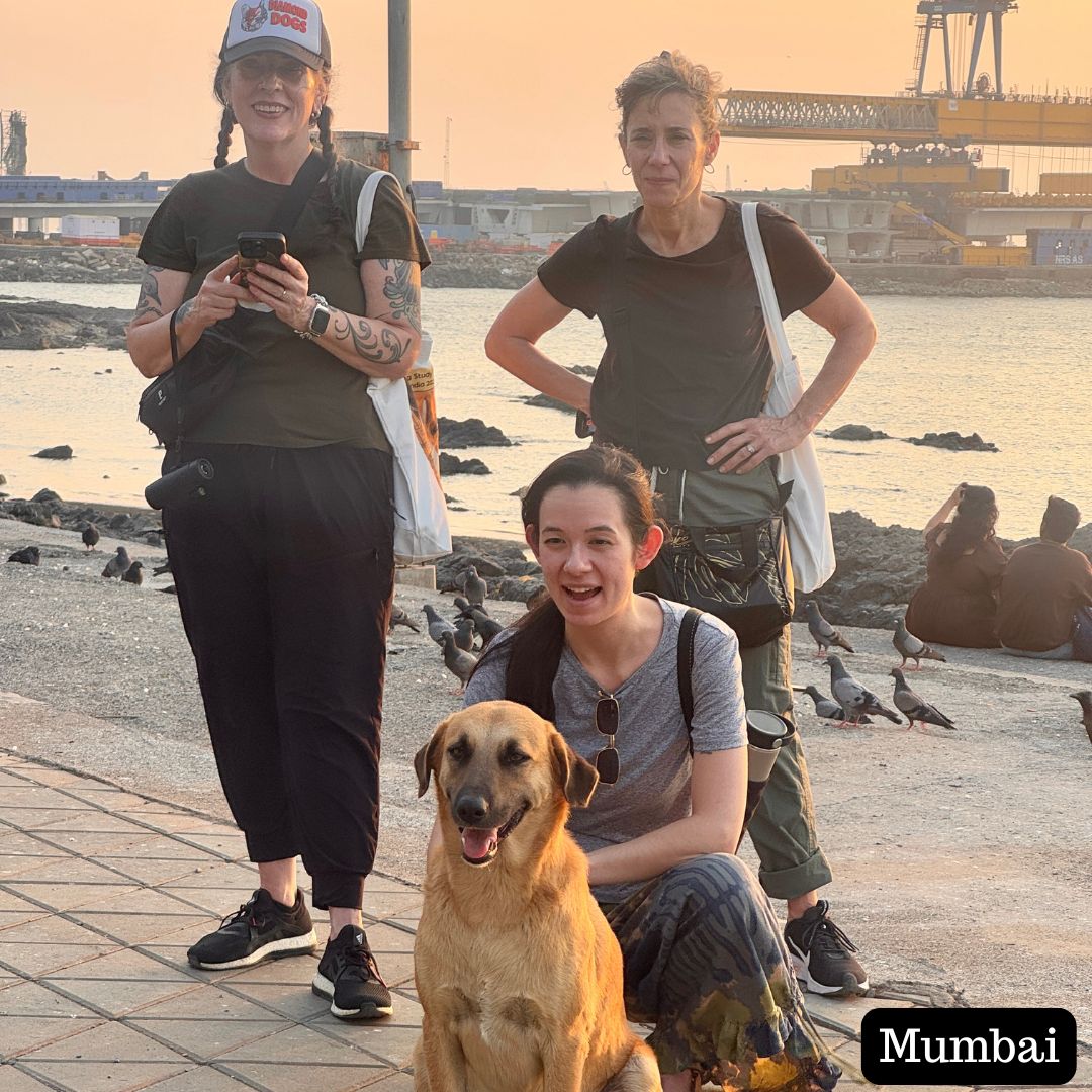 
                  
                    Three people with a dog on a waterfront promenade with a crane in the background, labeled 'Mumbai'.
                  
                