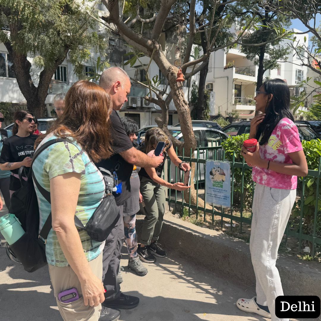 
                  
                    Group of people standing outdoors with a tree and buildings in the background, labeled 'Delhi'.
                  
                